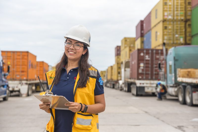 Female logistics coordinator at Tetakawi’s Mazatlán manufacturing community, standing in front of stacked shipping containers and freight trucks